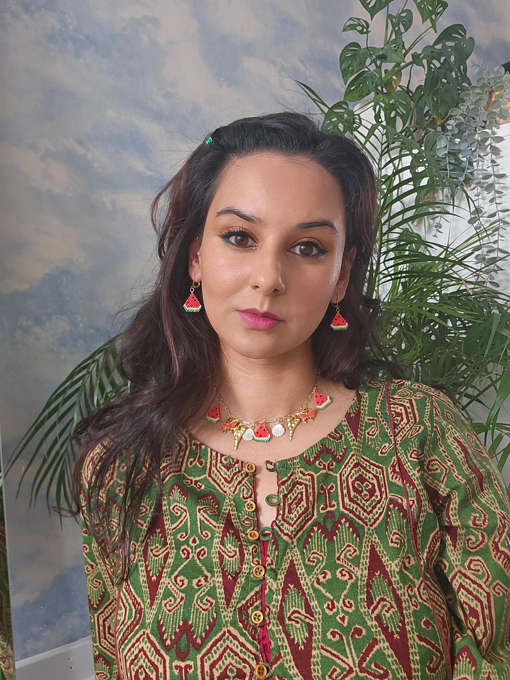 Woman wearing a patterned green and red outfit with jewelry, standing against a natural background with plants.